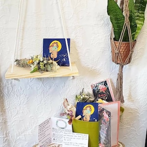 Puede incluir: Una estantería de madera con un colgador de macramé colgante con una planta verde. La estantería tiene una pequeña tarjeta con una mujer sosteniendo un globo terráqueo. Una mesa con un cubo verde lleno de flores secas, tarjetas y cristales. La mesa tiene una tarjeta con el texto "Por favor, recuerda que hay infinitas maneras de llevar tu luz única al mundo."