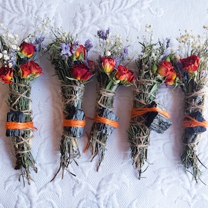May include: Five bundles of dried herbs and flowers tied with twine and a black stone. The bundles are decorated with orange string and include dried roses, rosemary, and other herbs.