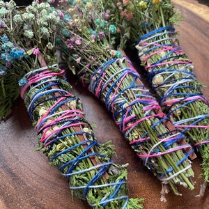 May include: Three bundles of dried herbs and flowers tied together with blue, pink, and white string. The bundles are arranged on a wooden surface.