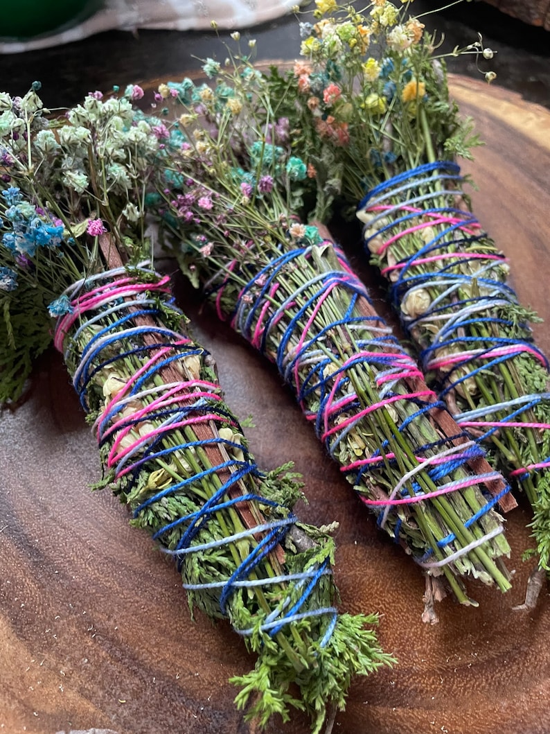 May include: Three bundles of dried herbs and flowers tied together with blue, pink, and white string. The bundles are arranged on a wooden surface.