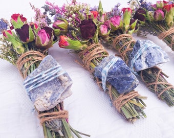 May include: Four bundles of dried flowers and herbs tied with twine and topped with a blue stone. The bundles are arranged in a row on a white surface.