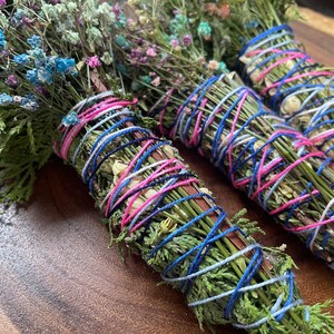 May include: Three bundles of dried herbs and flowers tied together with blue, pink, and white string. The bundles are arranged on a wooden surface.