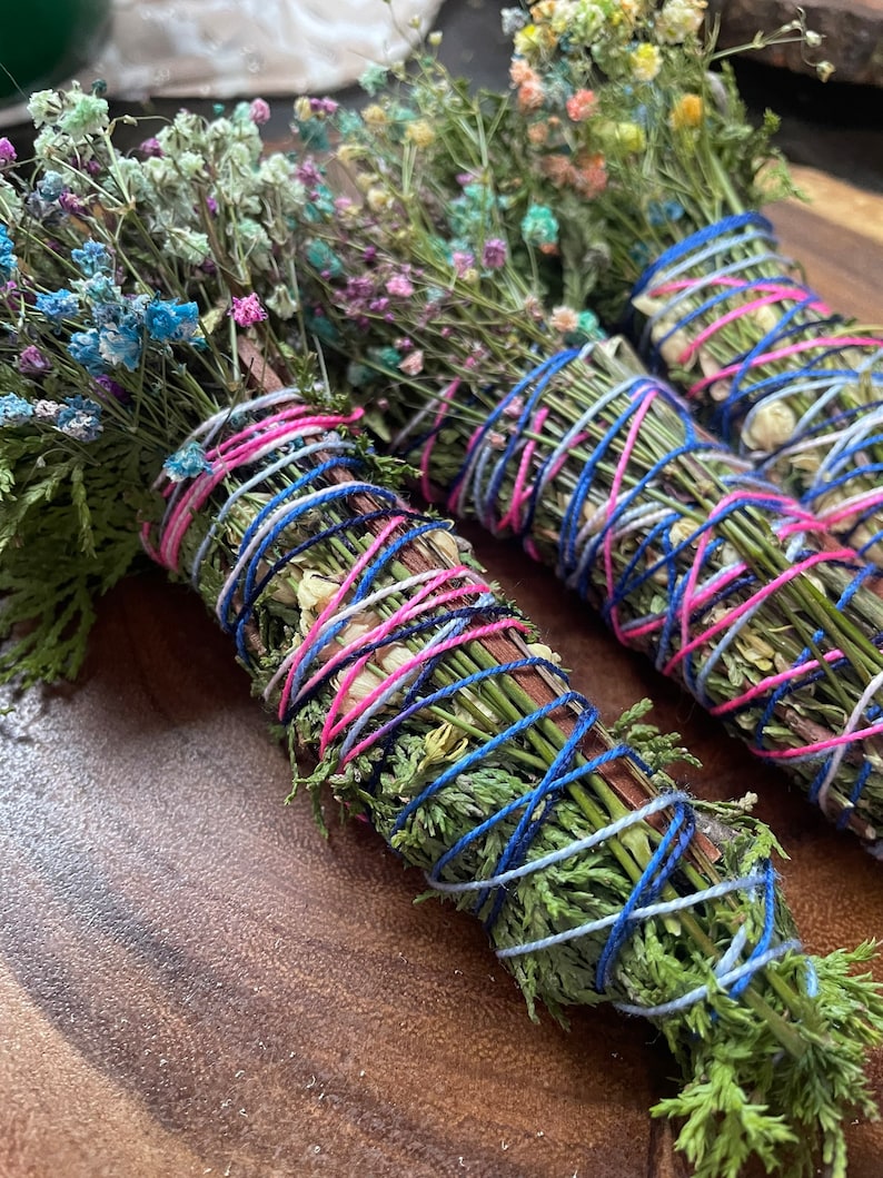 May include: Three bundles of dried herbs and flowers tied together with blue, pink, and white string. The bundles are arranged on a wooden surface.