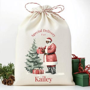 Könnte beinhalten: Cremefarbener Weihnachtsgeschenkebeutel mit rotem und weißem Kordelzug. Der Beutel zeigt eine Grafik von Santa Claus in rotem Anzug, einen Weihnachtsbaum und den Text "Special Delivery For Kailey."