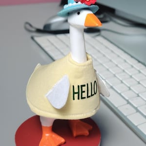May include: A whimsical goose figurine wearing a light blue hat adorned with red flowers and a yellow sweater with the word "HELLO" in green. The goose stands on a red base, with orange feet and beak.
