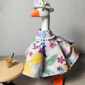 May include: A white goose figurine wearing a light gray hat with pastel flowers, a pearl necklace, and a dress featuring a colorful floral pattern in shades of purple, yellow, and green. The goose has an orange beak and orange feet.