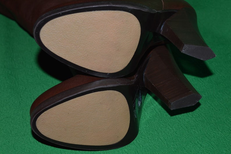 May include: Close-up of the bottom of two brown leather boots with a beige leather insole. The boots have a high heel.