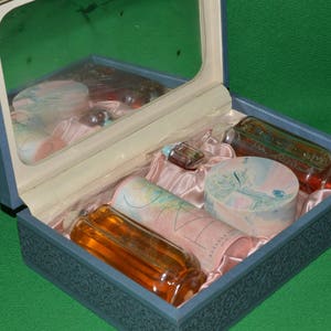 Pu&ograve; includere: Un astuccio da toilette blu con un coperchio a specchio, aperto per rivelare un set di articoli da toeletta. Il set include un flacone di profumo, un flacone di lozione, un flacone di olio da bagno, un sapone e una piccola scatola decorativa.