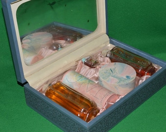 Puede incluir: Un estuche de tocador azul con tapa de espejo, abierto para revelar un conjunto de artículos de tocador. El conjunto incluye una botella de perfume, una botella de loción, una botella de aceite de baño, una pastilla de jabón y una pequeña caja decorativa.