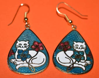 Puede incluir: Un par de pendientes colgantes con un fondo de esmalte azul turquesa y un diseño de gato blanco. El gato está rodeado de flores rosas y blancas.