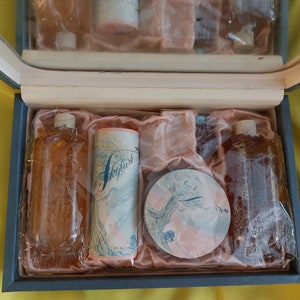 Pu&ograve; includere: Una scatola regalo blu con un rivestimento in raso rosa contiene un set di articoli da toeletta. Il set comprende due flaconi di vetro trasparente con un liquido marrone, un compatto rotondo rosa con un motivo floreale e un contenitore cilindrico rosa con un motivo floreale e la parola "Skylark" stampata su di esso.
