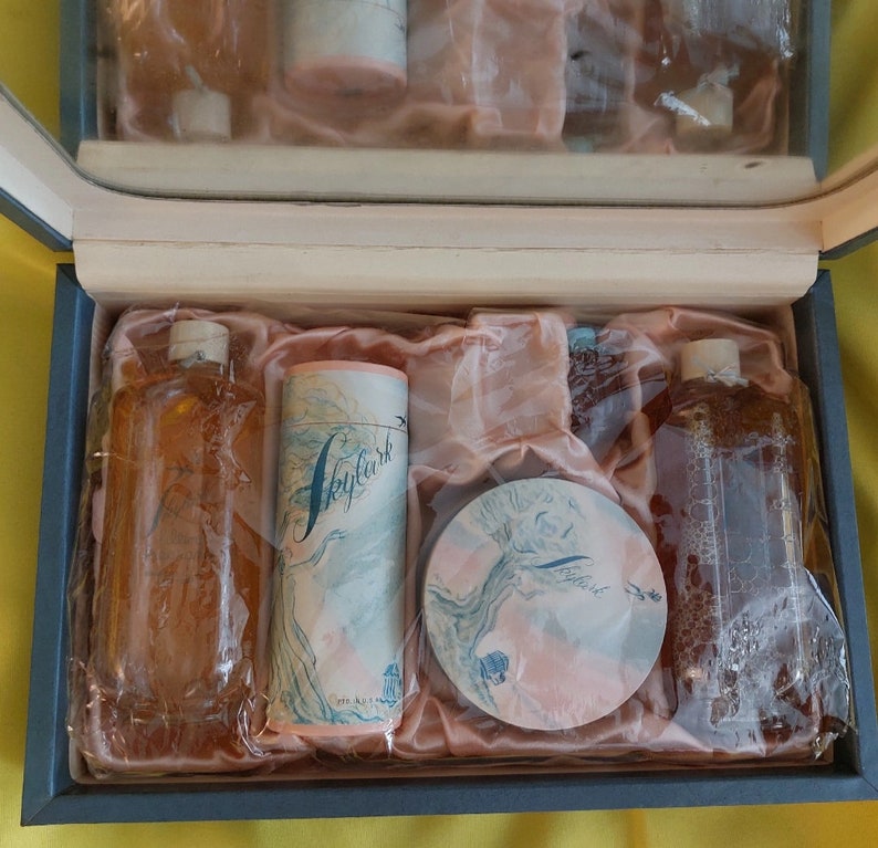 Pu&ograve; includere: Una scatola regalo blu con un rivestimento in raso rosa contiene un set di articoli da toeletta. Il set comprende due flaconi di vetro trasparente con un liquido marrone, un compatto rotondo rosa con un motivo floreale e un contenitore cilindrico rosa con un motivo floreale e la parola "Skylark" stampata su di esso.