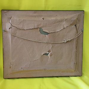 May include: The back of a framed picture with a brown paper backing. The paper is torn and ripped in several places. A string is attached to the frame for hanging.