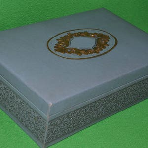 Pu&ograve; includere: Una scatola blu chiaro con un design decorativo dorato sulla parte superiore. La scatola ha un bordo decorato intorno ai bordi.