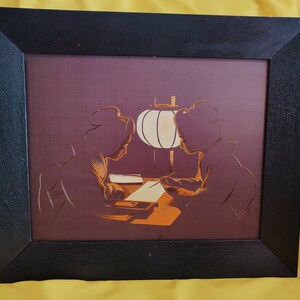 May include: A framed print depicting two women in traditional Japanese attire seated at a table with a paper lantern hanging overhead. The print is in shades of brown, orange, and white.