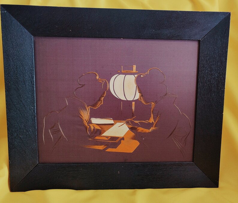 May include: A framed print depicting two women in traditional Japanese attire seated at a table with a paper lantern hanging overhead. The print is in shades of brown, orange, and white.