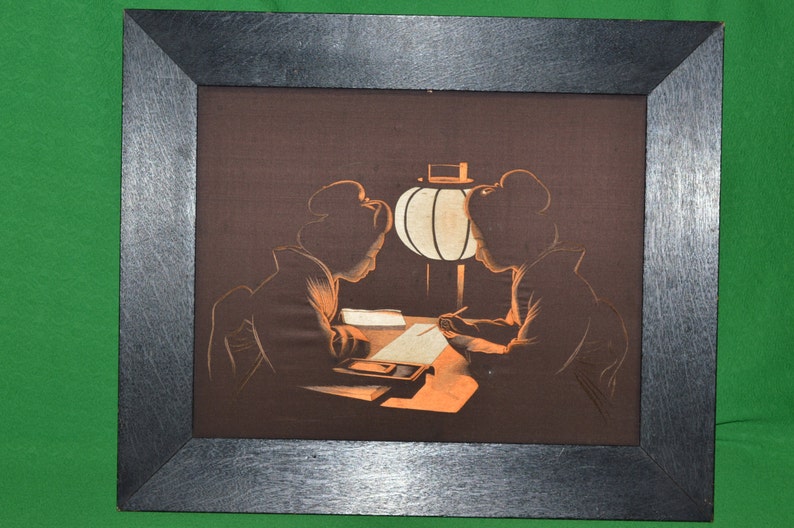 May include: A framed artwork depicting two figures in silhouette, seated at a table with a lamp. The figures are dressed in traditional Japanese clothing and appear to be writing or drawing. The artwork is set against a dark brown background.