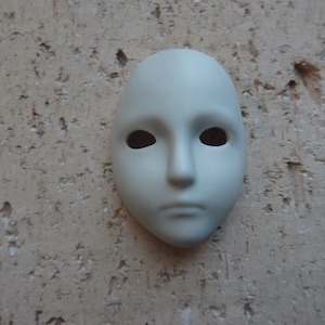 Peut inclure: Un masque blanc cassé avec des ouvertures ovales pour les yeux, un petit nez et une bouche légèrement tombante. Le masque est lisse et semble être en céramique. Le fond est une surface texturée, brun clair.