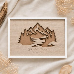 May include: A rectangular wooden wall art piece in a white frame featuring a laser-cut layered mountain and forest landscape. The design includes a central river flowing through trees and mountains with the engraved text Julia & Florian 09.08.2026.