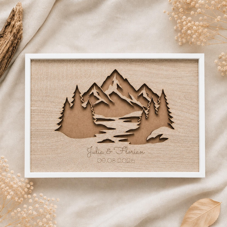 May include: A rectangular wooden wall art piece in a white frame featuring a laser-cut layered mountain and forest landscape. The design includes a central river flowing through trees and mountains with the engraved text Julia & Florian 09.08.2026.