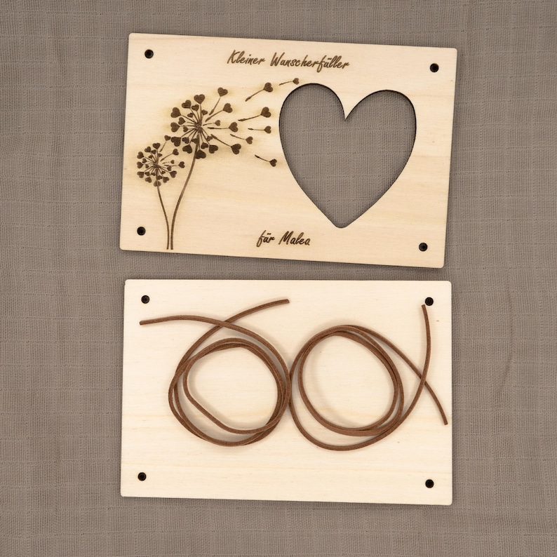 May include: Two wooden plaques with a heart-shaped cutout and a dandelion design. The top plaque reads "Kleiner Wunschf&auml;nger" and "f&uuml;r Malea". The bottom plaque has a brown cord wrapped around it.