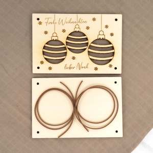 Pu&ograve; includere: Set di ornamenti natalizi in legno in due pezzi. La parte superiore presenta tre ornamenti appesi con strisce orizzontali, fiocchi di neve e il testo "Frohe Weihnachten lieber Noah". La parte inferiore ha due corde marroni.