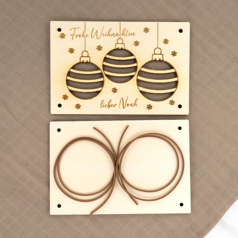 Pu&ograve; includere: Set di ornamenti natalizi in legno in due pezzi. La parte superiore presenta tre ornamenti appesi con strisce orizzontali, fiocchi di neve e il testo "Frohe Weihnachten lieber Noah". La parte inferiore ha due corde marroni.