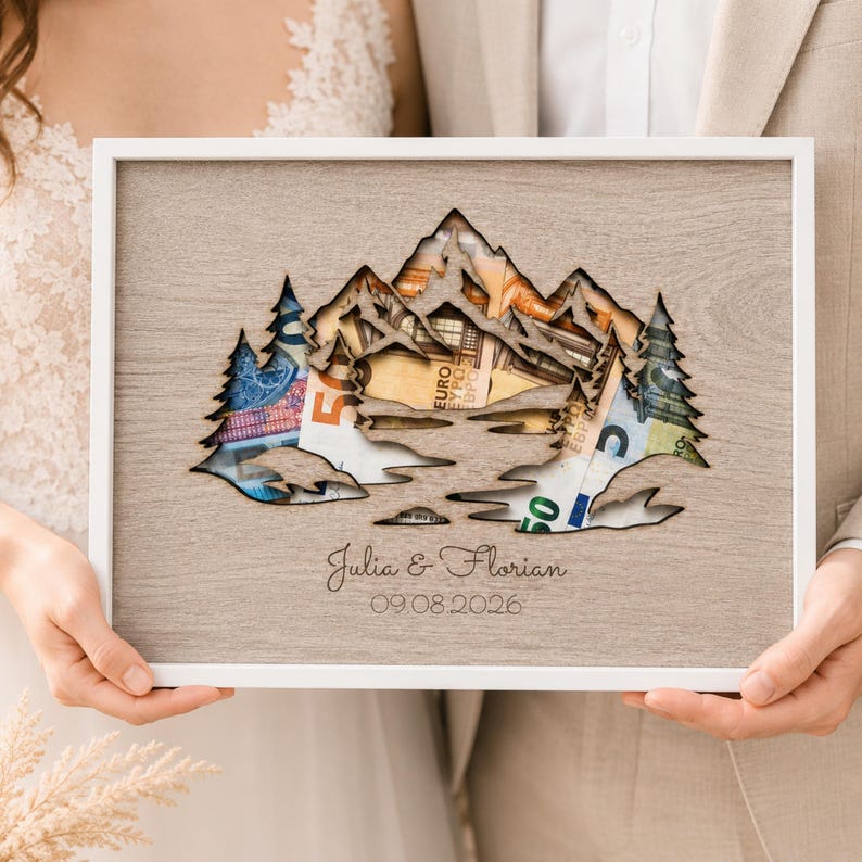 May include: A rectangular wooden frame containing a light brown wood-grain panel with a laser-cut mountain and pine tree silhouette. Behind the cutout, various Euro banknotes are visible. The text Julia & Florian and 09.08.2026 is engraved.