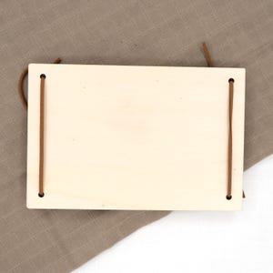 May include: A blank wooden sign with two brown leather straps attached to the top and bottom. The sign is on a brown fabric background.