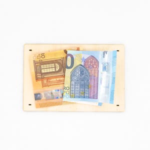 Pu&ograve; includere: Una tavola rettangolare in legno chiaro con quattro piccoli fori vicino agli angoli, su cui sono sovrapposte una banconota da 20 euro gialla e una da 20 euro blu. Le banconote mostrano illustrazioni architettoniche di finestre e archi.