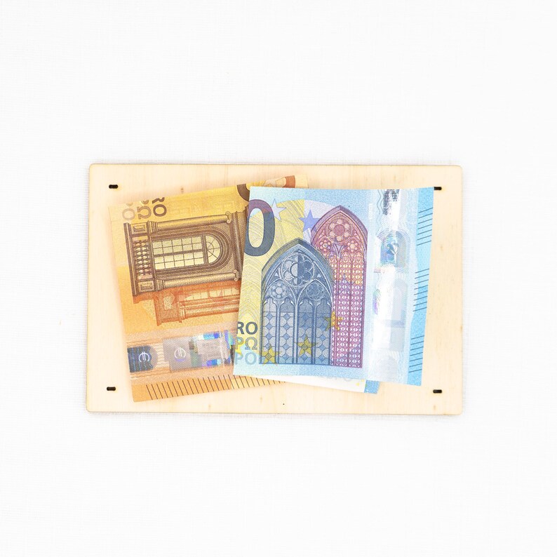 Pu&ograve; includere: Una tavola rettangolare in legno chiaro con quattro piccoli fori vicino agli angoli, su cui sono sovrapposte una banconota da 20 euro gialla e una da 20 euro blu. Le banconote mostrano illustrazioni architettoniche di finestre e archi.