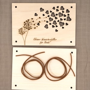 May include: A wooden card with a dandelion design and the text "Kleiner Wunscherf&uuml;ller f&uuml;r Noah!"  The card is accompanied by a brown cord.