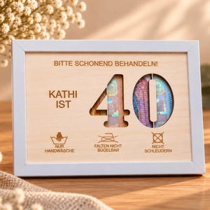 May include: A light-colored wooden frame containing a light wood panel with the text BITTE SCHONEND BEHANDELN!, KATHI IST, and the number 40 cut out. Below are laundry symbols with the text NUR HANDWÄSCHE, FALTEN NICHT BÜGELBAR, and NICHT SCHLEUDERN.