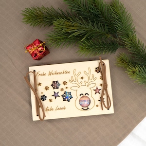 Pu&ograve; includere: Biglietto di Natale in legno con un design a renna, fiocchi di neve e stelle. La carta ha il testo "Frohe Weihnachten" e "liebe Leonie". Cordino marrone attaccato. Una piccola confezione regalo rossa e un ramo di pino sullo sfondo.