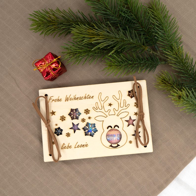 Pu&ograve; includere: Biglietto di Natale in legno con un design a renna, fiocchi di neve e stelle. La carta ha il testo "Frohe Weihnachten" e "liebe Leonie". Cordino marrone attaccato. Una piccola confezione regalo rossa e un ramo di pino sullo sfondo.