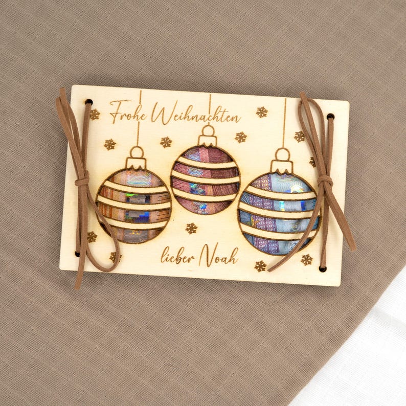 Pu&ograve; includere: Biglietto di Natale in legno con tre ornamenti appesi a strisce nei toni del marrone, viola e blu. La carta ha il testo "Frohe Weihnachten lieber Noah" ed &egrave; legata con un cordino in pelle scamosciata marrone. Decorazione festiva.