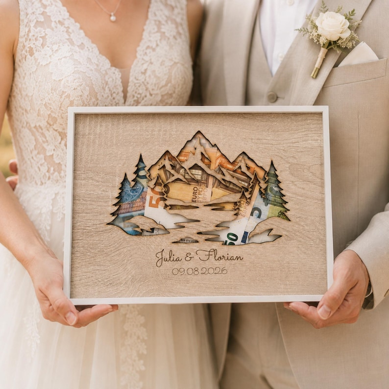 May include: A rectangular wooden frame featuring a laser-cut mountain and pine tree silhouette. The cutout reveals Euro banknotes placed behind the wood. The front surface is engraved with the names Julia & Florian and the date 09.08.2026.