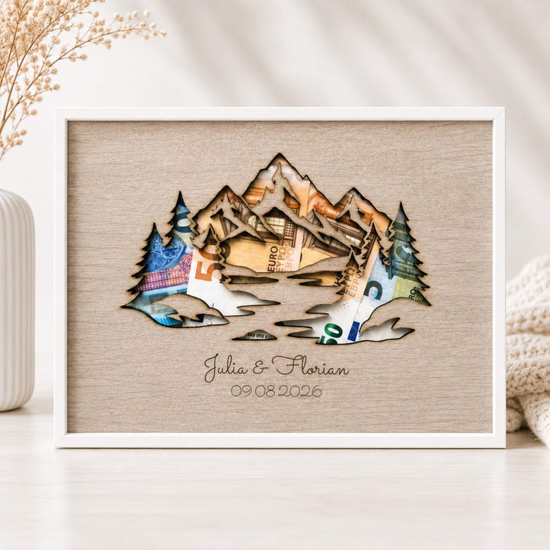 May include: A rectangular wooden frame containing a light brown panel with a laser-cut mountain and pine tree silhouette. Behind the cutout, various Euro banknotes are visible. The text Julia & Florian and 09 08 2026 is engraved below.