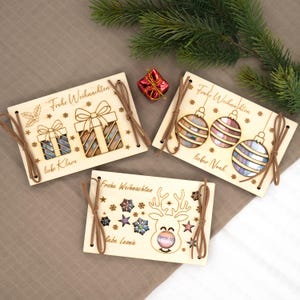 Pu&ograve; includere: Tre biglietti di Natale in legno con disegni tagliati al laser. Un biglietto presenta confezioni regalo, un altro ornamenti e il terzo una renna. Ogni biglietto ha uno spago marrone e le parole "Frohe Weihnachten" e un nome. Una piccola confezione regalo rossa e verde sono sullo sfondo.