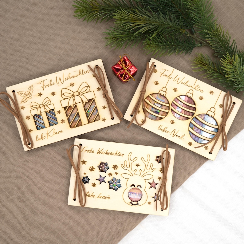 Pu&ograve; includere: Tre biglietti di Natale in legno con disegni tagliati al laser. Un biglietto presenta confezioni regalo, un altro ornamenti e il terzo una renna. Ogni biglietto ha uno spago marrone e le parole "Frohe Weihnachten" e un nome. Una piccola confezione regalo rossa e verde sono sullo sfondo.