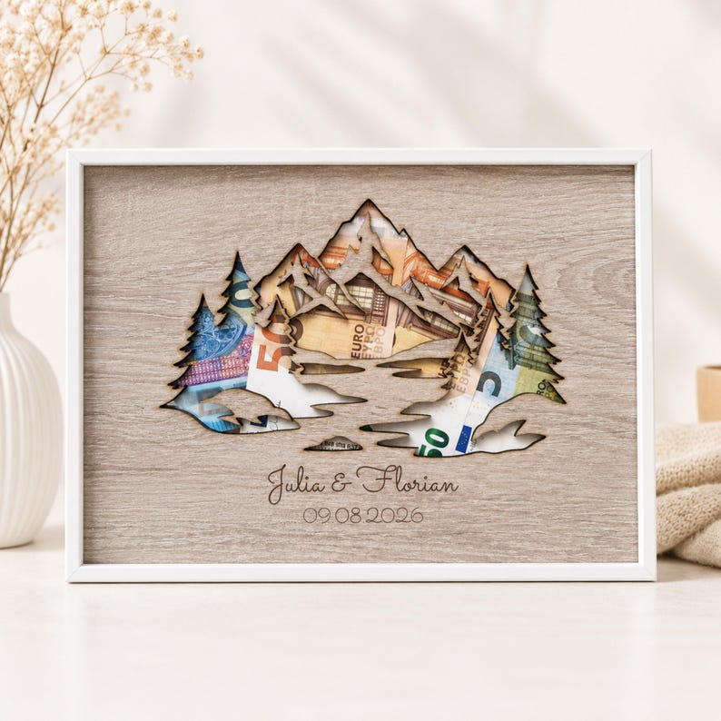 May include: A rectangular wooden frame featuring a laser-cut mountain and pine tree silhouette. Behind the cutout, colorful Euro banknotes are visible. The frame is engraved with the names Julia & Florian and the date 09.08.2026 in a script font.