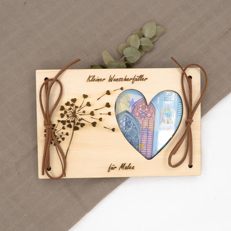 May include: A wooden box with a heart-shaped cutout, revealing money inside. The box is decorated with a dandelion and the text "Kleiner Wanscherfaller f&uuml;r Malea".