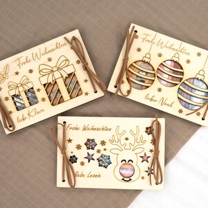 Pu&ograve; includere: Tre biglietti di Natale in legno con disegni tagliati al laser. Un biglietto presenta confezioni regalo, un altro ornamenti e l'ultimo mostra una renna. Ogni biglietto ha un cordino marrone e le parole "Frohe Weihnachten" e un nome.