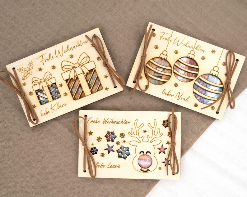 Pu&ograve; includere: Tre biglietti di Natale in legno con disegni tagliati al laser. Un biglietto presenta confezioni regalo, un altro ornamenti e l'ultimo mostra una renna. Ogni biglietto ha un cordino marrone e le parole "Frohe Weihnachten" e un nome.