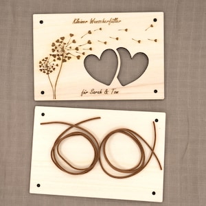 May include: A wooden craft kit with two pieces. The top piece features a dandelion with hearts cut out and the text "Kleiner Wunschf&auml;nger" and "f&uuml;r Sarah & Tom". The bottom piece has two brown cords.