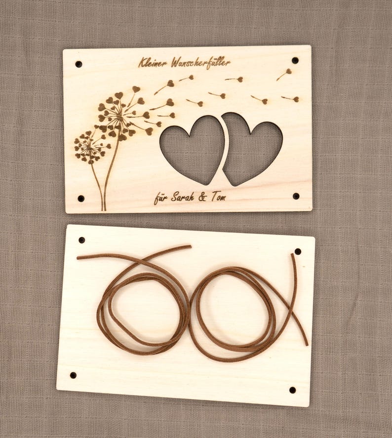 May include: A wooden craft kit with two pieces. The top piece features a dandelion with hearts cut out and the text "Kleiner Wunschf&auml;nger" and "f&uuml;r Sarah & Tom". The bottom piece has two brown cords.