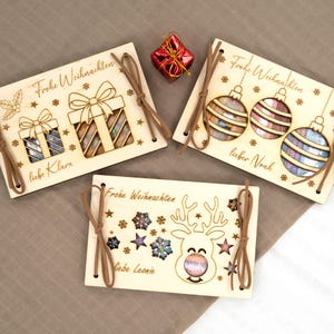 Pu&ograve; includere: Ornamenti natalizi in legno con disegni tagliati al laser. Uno presenta regali, un altro ornamenti e l'ultimo una renna. Ognuno ha un cordino in pelle per appenderlo e il testo tedesco "Frohe Weihnachten" e nomi come "Klara" e "Noah".