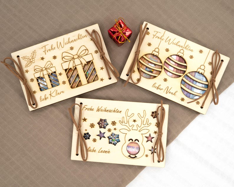 Pu&ograve; includere: Ornamenti natalizi in legno con disegni tagliati al laser. Uno presenta regali, un altro ornamenti e l'ultimo una renna. Ognuno ha un cordino in pelle per appenderlo e il testo tedesco "Frohe Weihnachten" e nomi come "Klara" e "Noah".