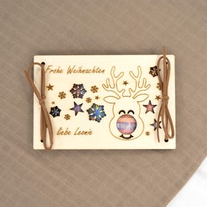 Pu&ograve; includere: Ornamento natalizio in legno con un design a renna, stelle e fiocchi di neve. Le parole "Frohe Weihnachten" e "liebe Leonie" sono incise nel legno. Corde in pelle marrone per appenderlo.