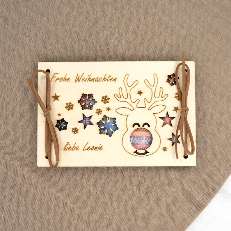 Pu&ograve; includere: Ornamento natalizio in legno con un design a renna, stelle e fiocchi di neve. Le parole "Frohe Weihnachten" e "liebe Leonie" sono incise nel legno. Corde in pelle marrone per appenderlo.